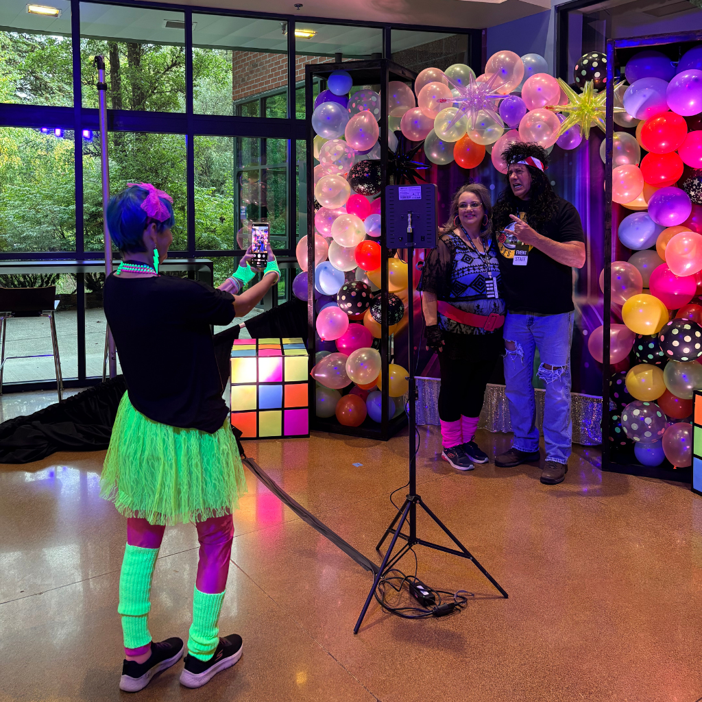 Volunteers at an 80s themed photo booth. One volunteer is taking the picture for 2 other volunteers.